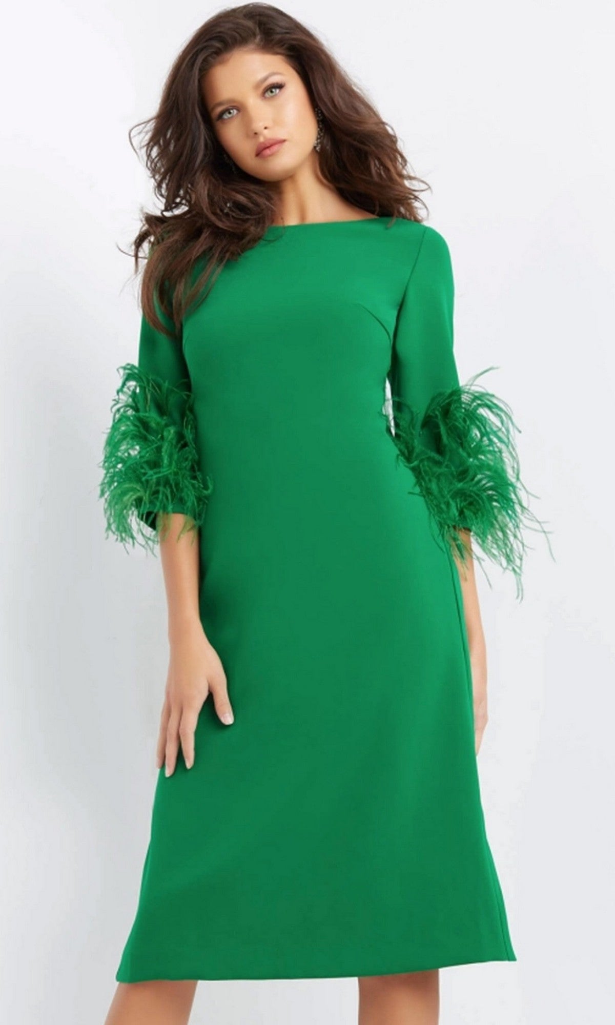 A woman wearing the Jovani 07341 short wedding guest dress, featuring a knee-length green design with elegant feathered detailing on the sleeves, stands against a plain light background.
