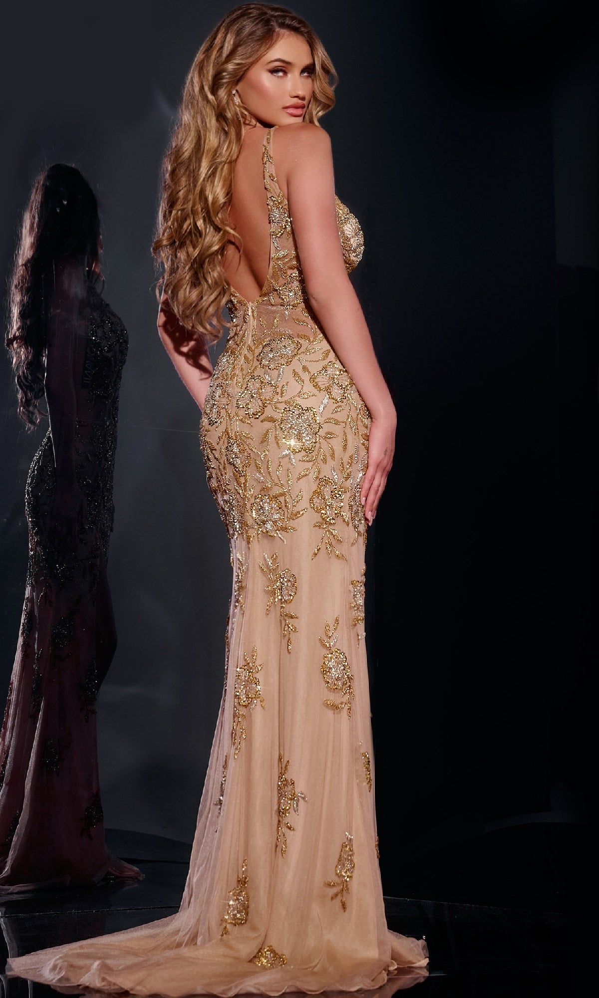 A woman with long wavy hair wears the backless gold Jovani 07355 prom dress, featuring intricate embroidery, as she stands with her back to the camera in a softly lit room, her reflection shimmering in a nearby mirror.