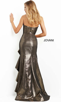 A woman stands facing a white wall, her back elegantly visible in the strapless metallic Long Formal Dress: Jovani 07582. The mermaid gown flares at the bottom with cascading ruffle accents, creating a stunning silhouette. “JOVANI” is written beside her.