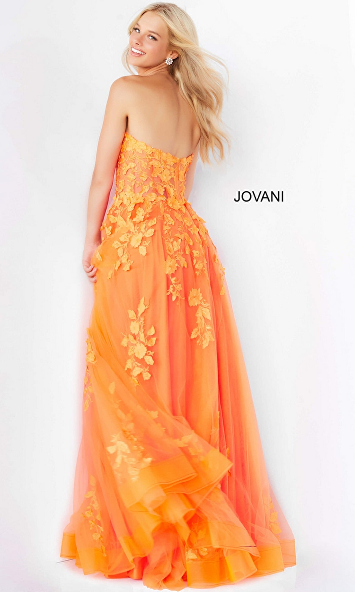 A woman wearing the Jovani Strapless Long Lace A-Line Prom Dress 07901, a floor-length orange gown with yellow floral appliqué, stands smiling over her shoulder. The word 
