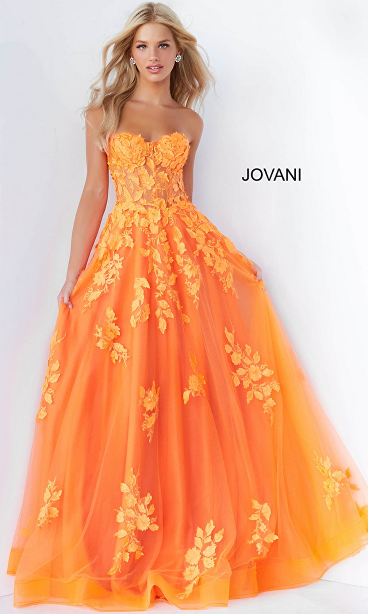 A woman stands against a white background wearing the Jovani Strapless Long Lace A-Line Prom Dress 07901 in orange, detailed with yellow floral appliqués. The word 