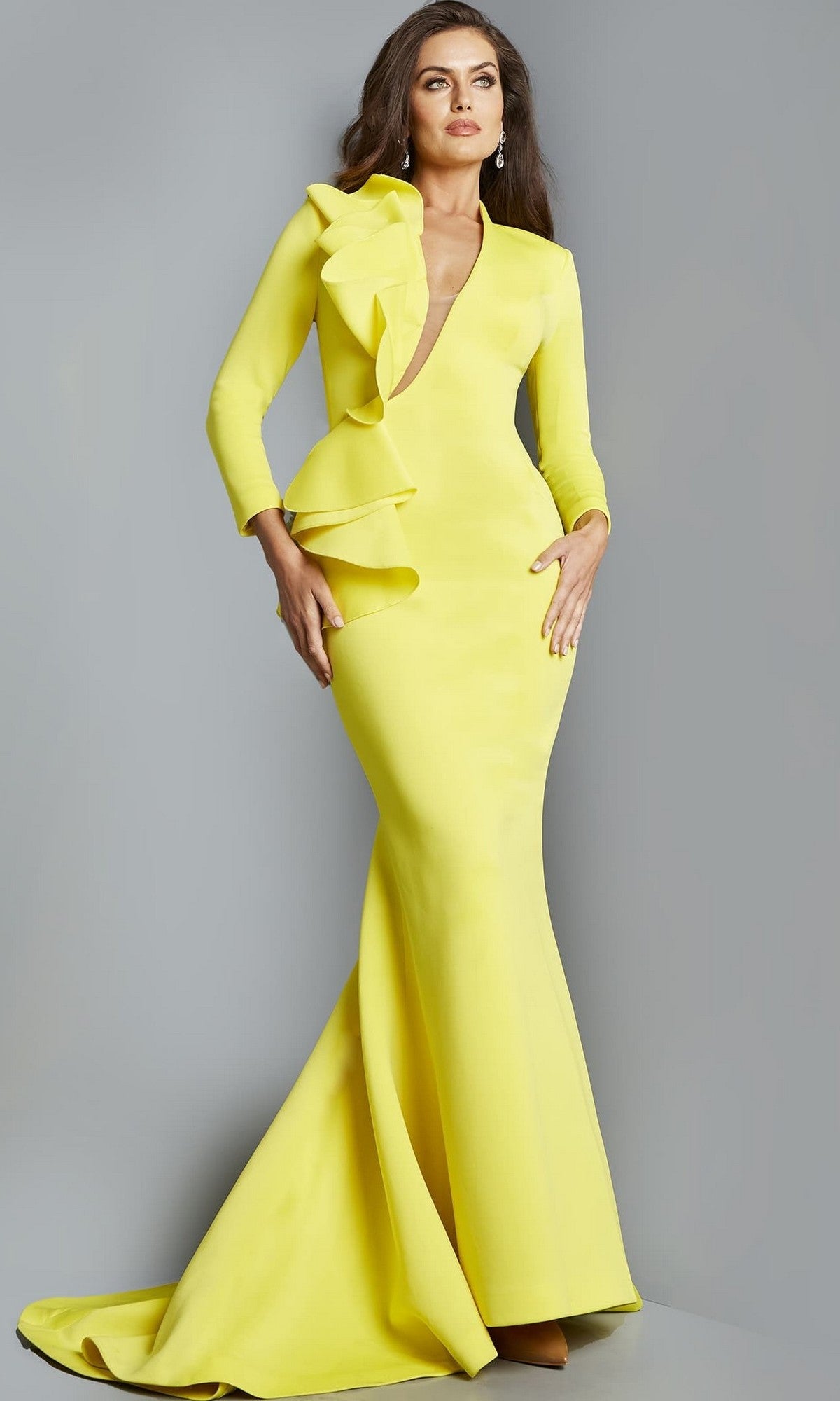 A woman stands against a plain gray background wearing the Jovani 07934 long formal dress in yellow—a striking, form-fitting gown with long sleeves, ruffle detail on one shoulder, and a deep V neckline. This elegant design makes it an ideal choice for a mother-of-the-bride seeking a stunning designer look.
