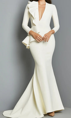 A woman stands against a plain gray background wearing the Long Formal Dress: Jovani 07934 in ivory, featuring a long, fitted silhouette with long sleeves, a deep V neckline, and ruffle detailing on one side.