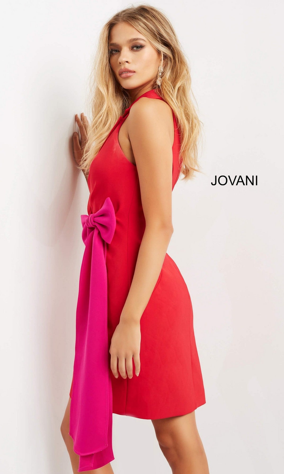 A woman in the Jovani 07961 Short Party Dress, a vibrant red sleeveless dress with a large pink bow at the waist, stands against a white wall. The brand name 