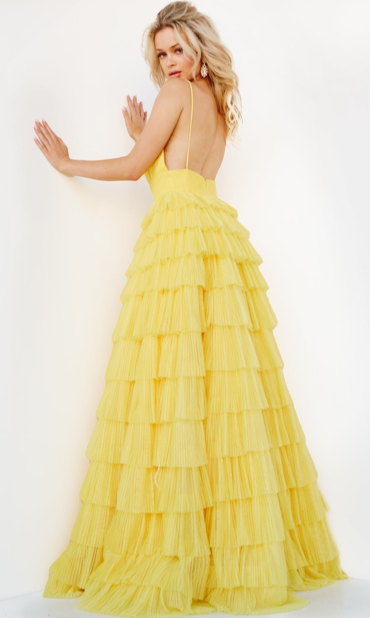 A woman poses with her back to the camera in the Jovani Open-Back Long Tiered Prom Ball Gown 08480, a yellow spaghetti-strap dress, against a white background.