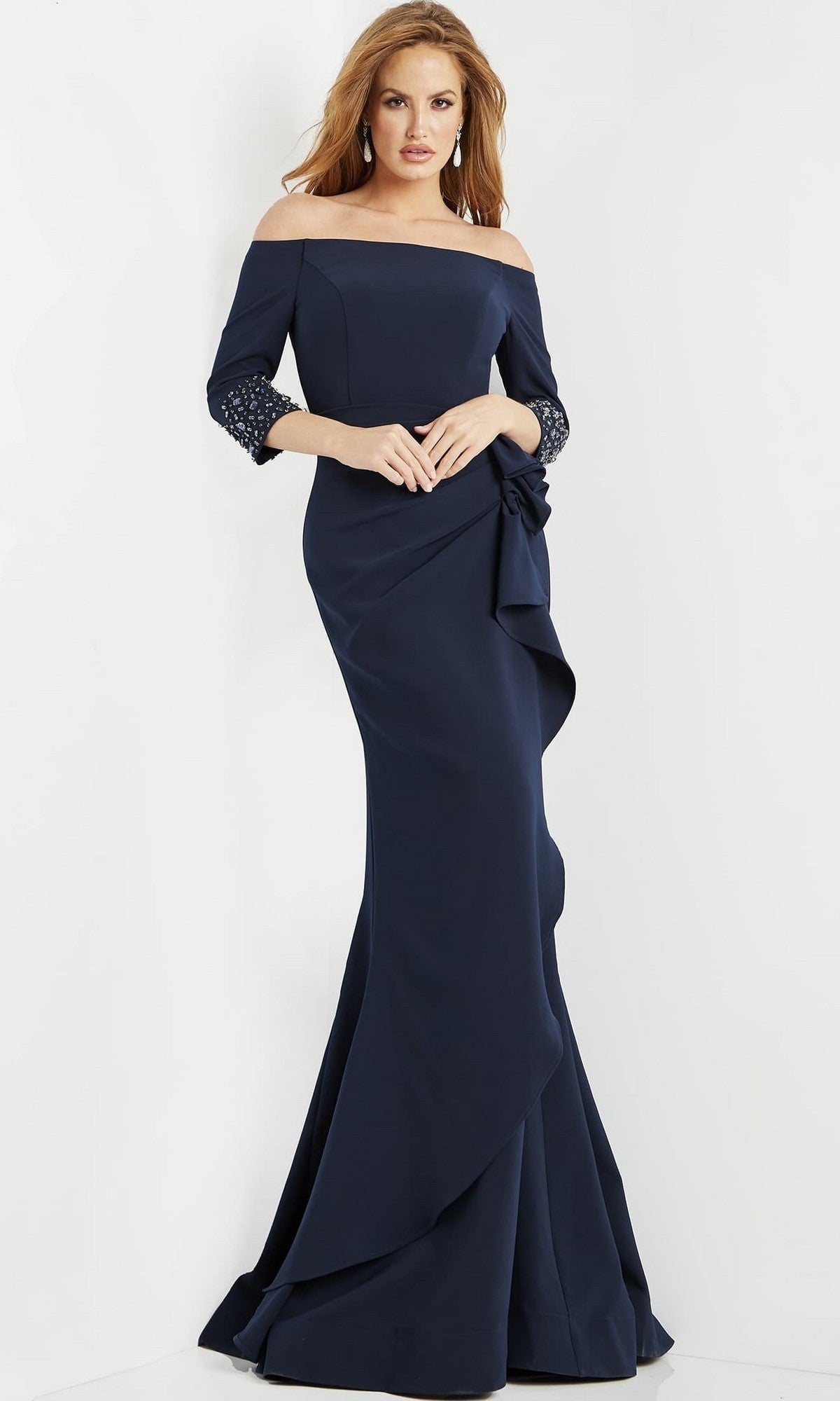 A woman stands against a white background wearing the Long Formal Dress: Jovani 08699—an elegant, off-shoulder navy blue gown featuring lace-trimmed sleeves and a floral detail at the waist.