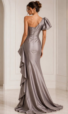 A woman is standing in a white room, photographed from the back, wearing the Jovani 09201 long formal dress—a fitted metallic silver gown with a one-shoulder design, ruffled accent, and elegant train.
