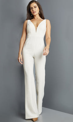 A woman stands against a gray wall wearing the Jovani 09479 Long Jumpsuit—a sleeveless, white, wide-leg formal jumpsuit with a deep V-neckline—paired with beige high heels.
