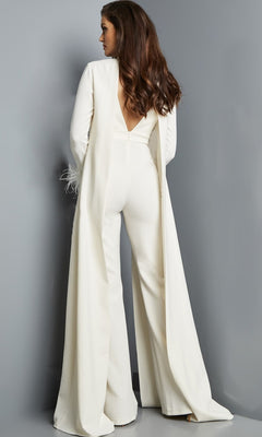 A woman stands facing away against a gray background, wearing the Jovani 09479 long jumpsuit in ivory crepe, featuring sleeveless styling, wide legs, and a dramatic cape-like overlay.
