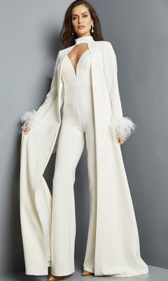 An elegant woman wears the ivory Long Jumpsuit: Jovani 09479, featuring a formal deep V-neckline and a long matching cape with feathered cuffs, standing against a plain gray background.