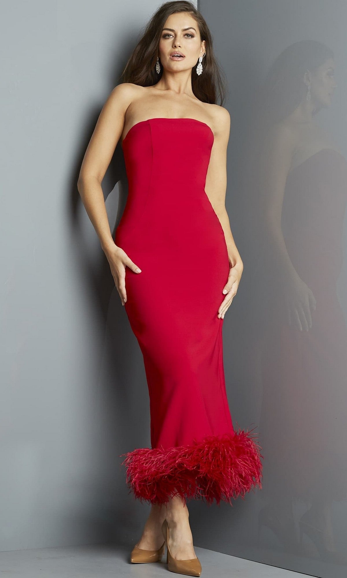 A woman stands against a gray wall wearing the Jovani 09630 Long Formal Dress in red, a strapless and form-fitting midi designer piece featuring a dramatic feather hemline at the bottom, complemented by tan heels and drop earrings.
