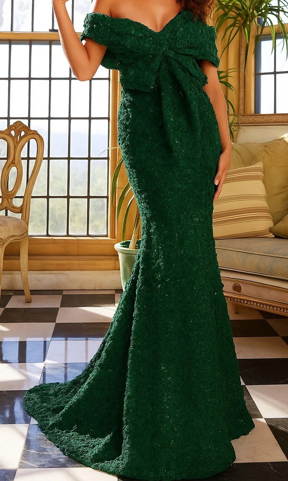 A woman stands in a room with checkered flooring and large windows, wearing the Jovani 09768 long formal dress in emerald. The off-the-shoulder, floor-length gown is adorned with a large front bow and features a graceful mermaid silhouette that elegantly highlights her figure.