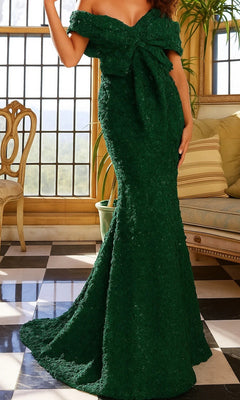 A woman stands in a room with checkered flooring and large windows, wearing the Jovani 09768 long formal dress in emerald. The off-the-shoulder, floor-length gown is adorned with a large front bow and features a graceful mermaid silhouette that elegantly highlights her figure.