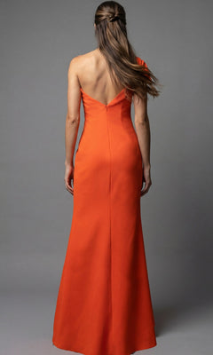 A woman wearing the Jovani 09961 long formal dress in orange stands facing away, showcasing its V-shaped back and one-shoulder design, with straight hair against a plain gray background.
