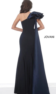 A woman stands with her back to the camera, wearing the Jovani 1008—a long formal designer dress featuring a black, one-shoulder, floor-length gown adorned with a large dark blue shoulder bow and trailing sash. The elegant sheath silhouette of the dress accentuates her figure and exudes sophistication.