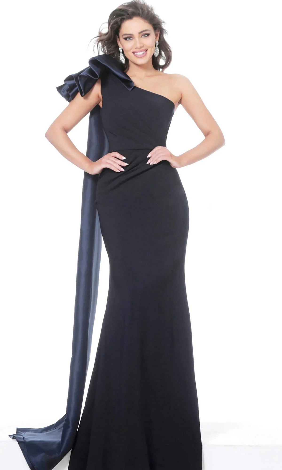 A woman stands smiling in the Jovani 1008 long formal dress—a navy, floor-length designer gown featuring an elegant sheath silhouette, one-shoulder design with a large ruffle detail and a long sash draping down—highlighted against a plain white background.