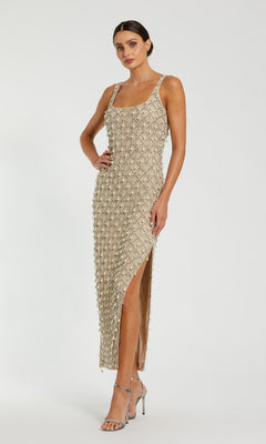 A woman stands against a white background wearing the Mac Duggal 10084 long formal dress in nude silver, featuring sleeveless styling, pearl embellishments, a side slit, and paired with silver high-heeled sandals.