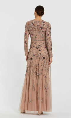 A woman stands with her back to the camera, wearing the Mac Duggal 10093 Long Sleeve Beaded Formal Dress in a sheer nude shade with intricate floral embroidery and beading, her hair styled neatly in a bun.