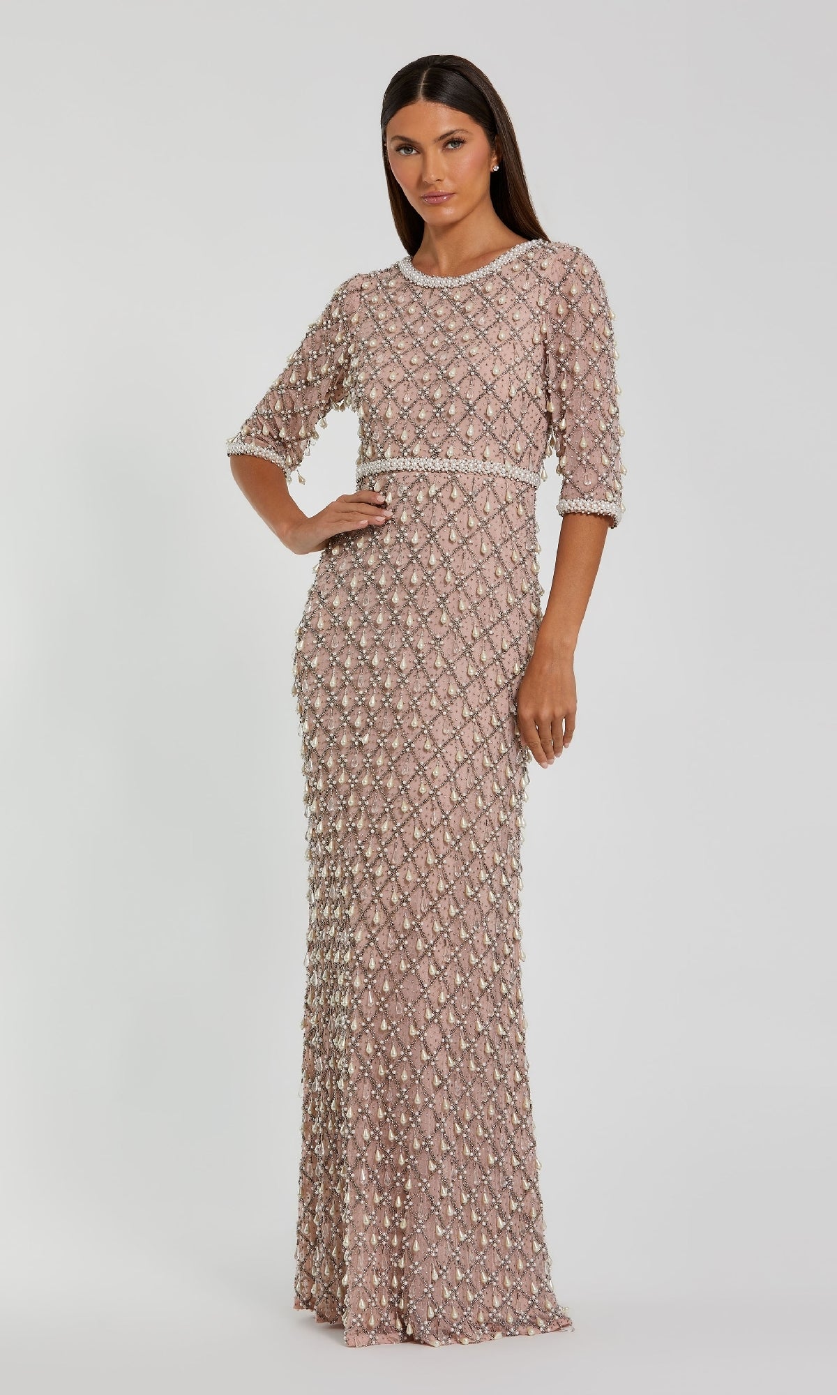 A woman wears the Beaded Long Pink Formal Dress: Mac Duggal 10099, featuring pearl embellishments, half sleeves, a fitted waist, and a floor-length silhouette, standing against a plain light background.