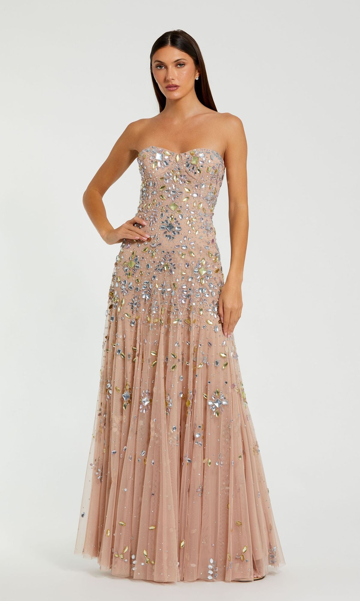 A woman poses gracefully against a white background in the Mac Duggal 10100, a strapless light pink prom gown adorned with silver and gold sequins.