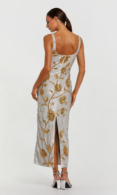 A woman is seen from behind wearing the Mac Duggal 10119 long formal dress in silver, featuring gold floral designs, a back slit, and paired with silver high-heeled sandals against a plain background.