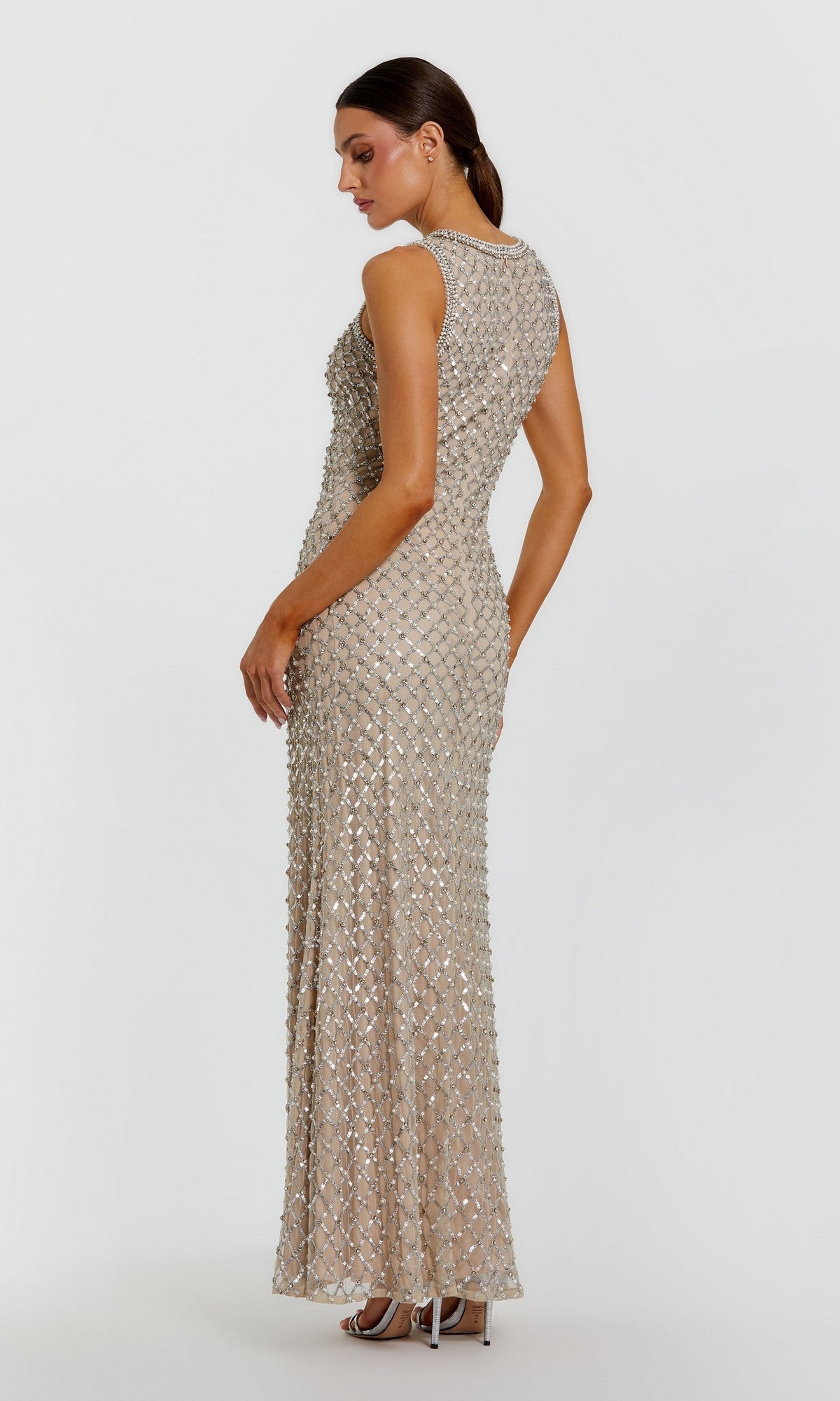 A woman stands facing away against a plain white background, wearing the Mac Duggal 10129 Long Formal Dress—a sleeveless, floor-length gown in silver-nude that features a fitted silhouette and intricate geometric beaded detailing.