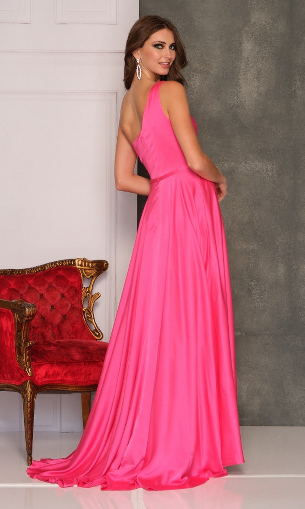 A woman wearing the Long Formal Dress A10524 by Dave and Johnny, a vibrant pink floor-length A-line gown, stands beside a red ornate chair, glancing over her shoulder with a smile.