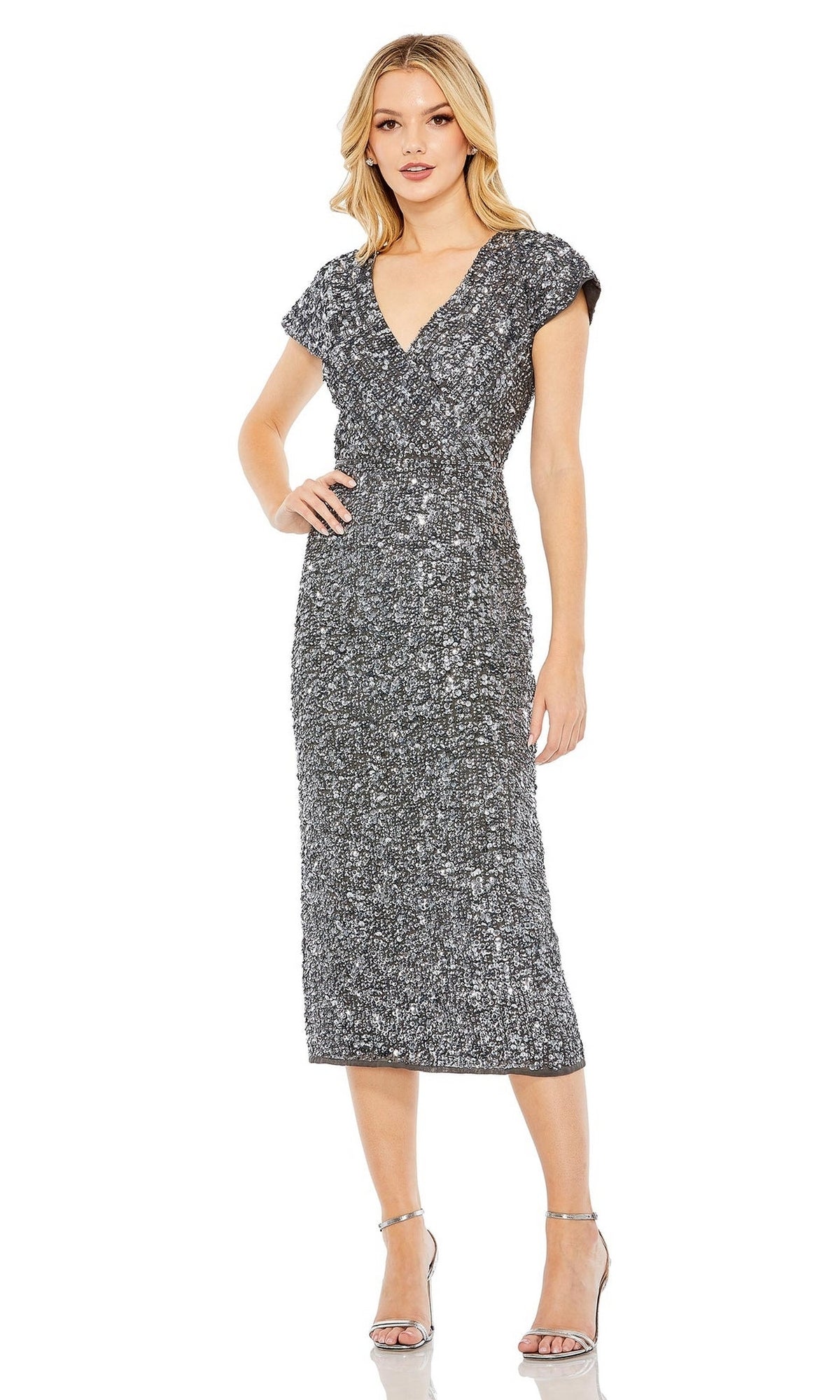 A woman stands against a plain white background wearing the Mac Duggal 10766 midi sequin wedding guest dress with cap sleeves, a V-neckline, and silver high-heeled sandals.