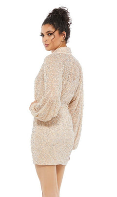 A woman with curly hair in a bun models the Short Wedding Guest Dress 10790 by Mac Duggal—a nude mini sequin dress with sheer long sleeves—standing with her back turned and looking over her shoulder against a white background.
