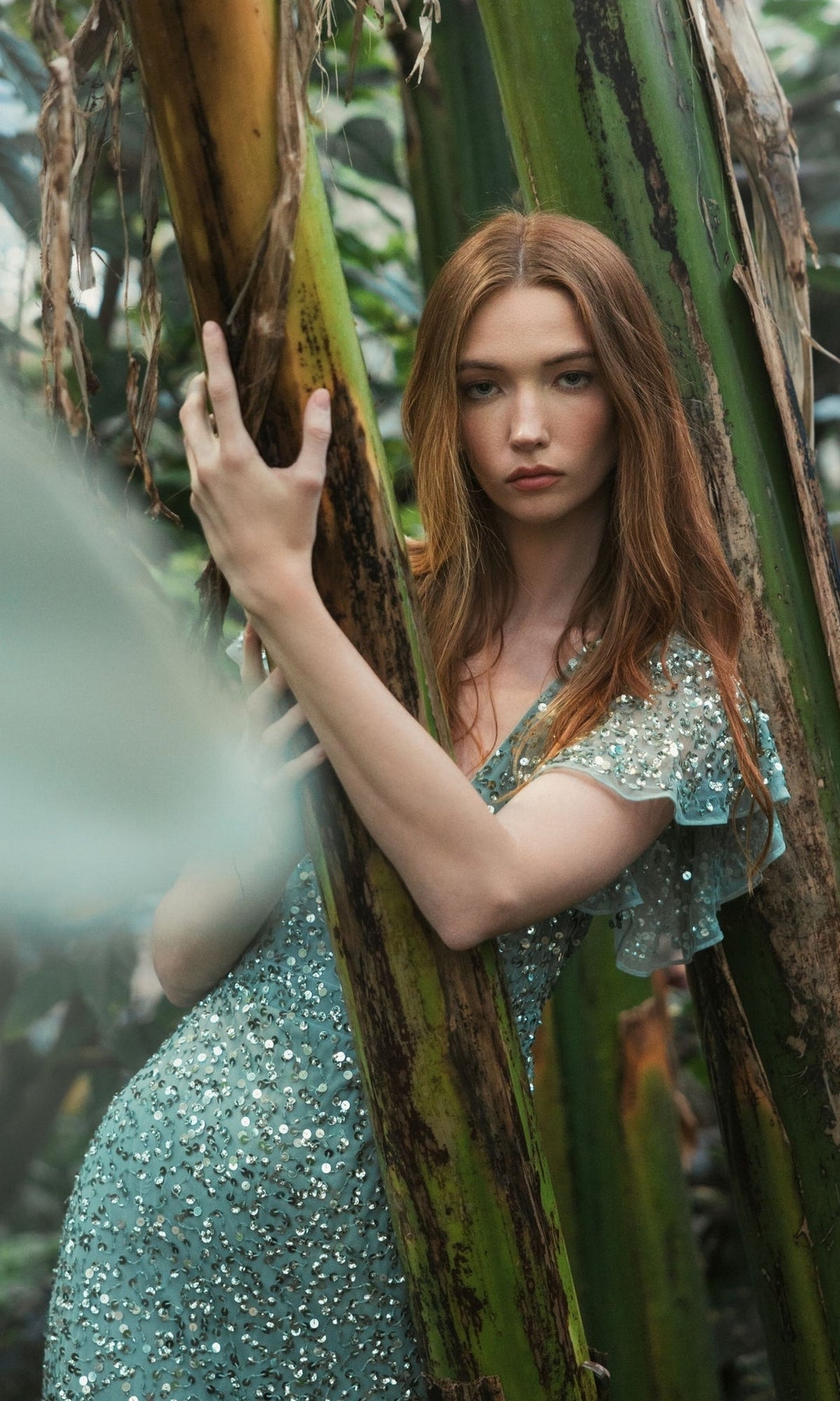 A woman with long red hair wears the Short Wedding Guest Dress 10801 by Mac Duggal, standing among tall green plants and holding a tree trunk as she looks at the camera.