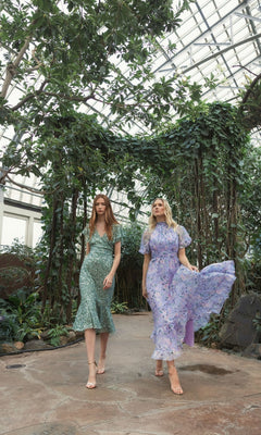 Two women wearing flowy, floral print dresses and one in the Mac Duggal Short Wedding Guest Dress 10801 walk along a garden path inside a greenhouse with lush greenery and a glass ceiling.