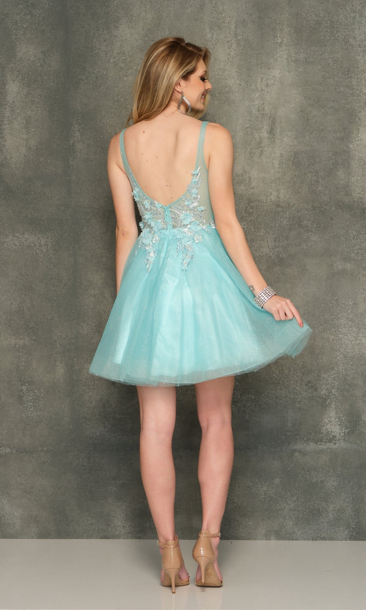 A woman in the Sheer-Bodice Short Homecoming Dress—light blue, a-line with floral appliques and a deep V back—stands against a gray textured background, facing away from the camera.