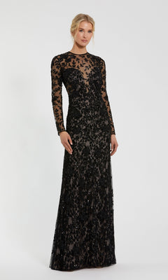 A woman wears the Mac Duggal 10821 long black beaded formal dress, featuring sheer fabric and intricate black floral embroidery, standing against a white backdrop.