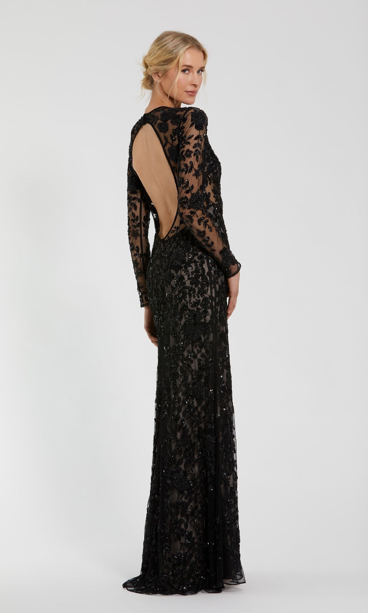 A woman stands against a plain background, looking over her shoulder in the Mac Duggal 10821 long black beaded formal dress with lace sleeves and an open back.