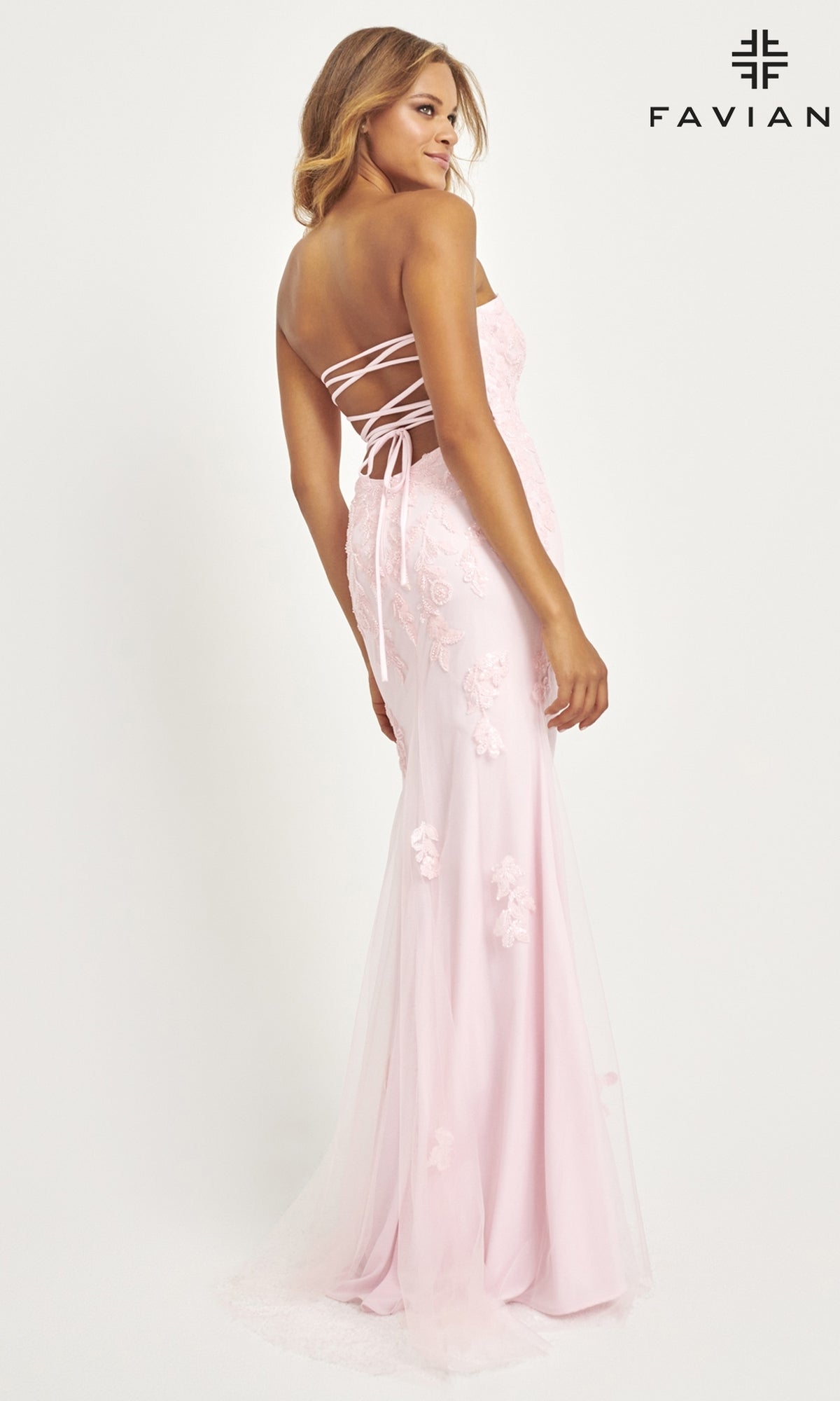 A woman wearing the Faviana Strapless Long Beaded Prom Dress 11004 in light pink, featuring a lace-up back and floral appliqué, stands looking over her shoulder against a plain white background with the Faviana logo in the top right.
