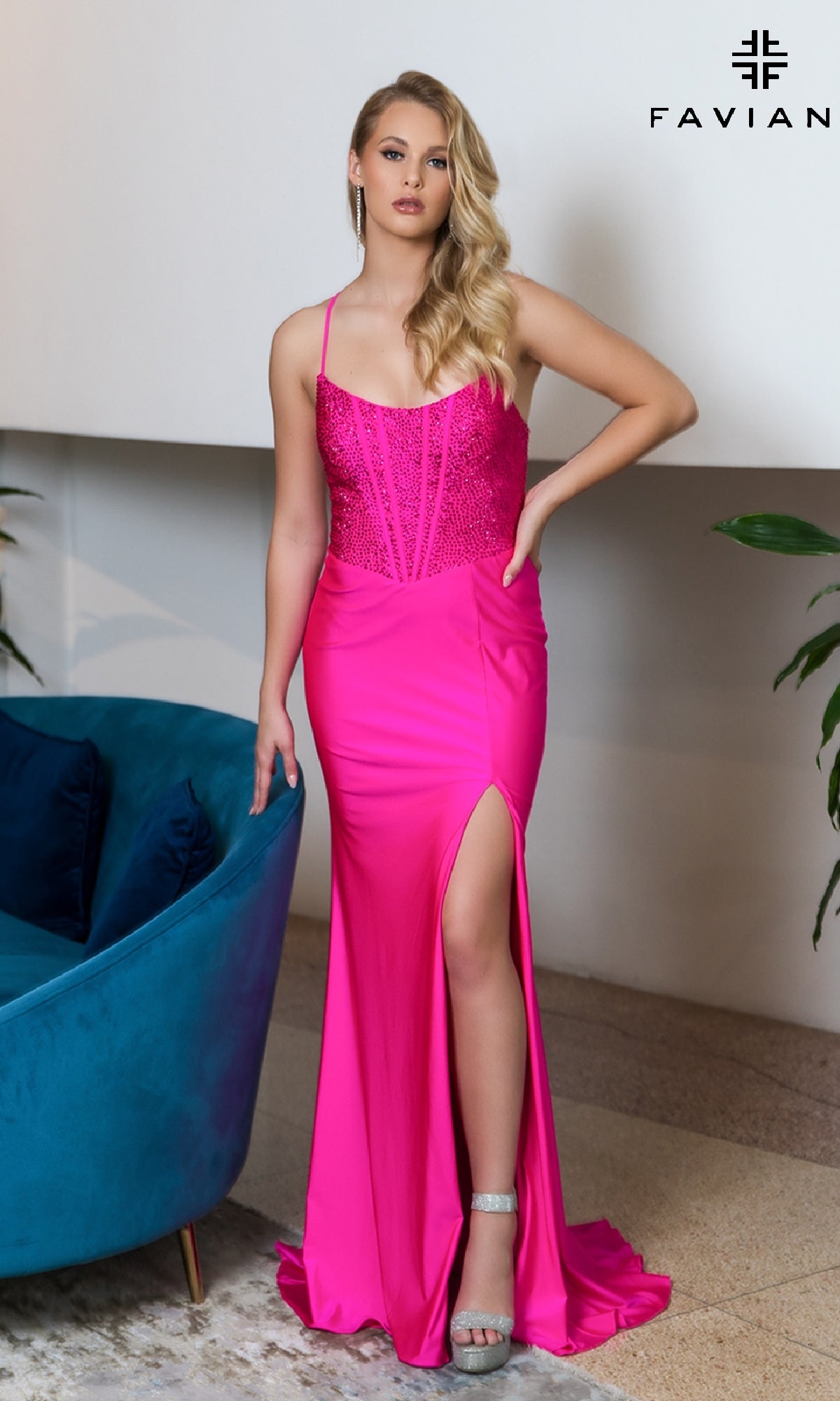 A woman with long blonde hair stands indoors by a blue chair wearing the Beaded-Corset-Bodice Faviana Long Prom Dress 11011 in bright pink, featuring a beaded bodice, thigh-high slit, and silver high heels.