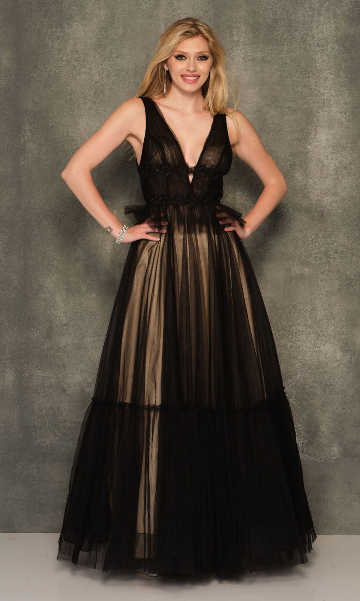 A woman stands before a gray backdrop in the Dave and Johnny Long Black Prom Ball Gown 11026, a sleeveless, floor-length A-line dress with a sheer bodice and relaxed tulle skirt.