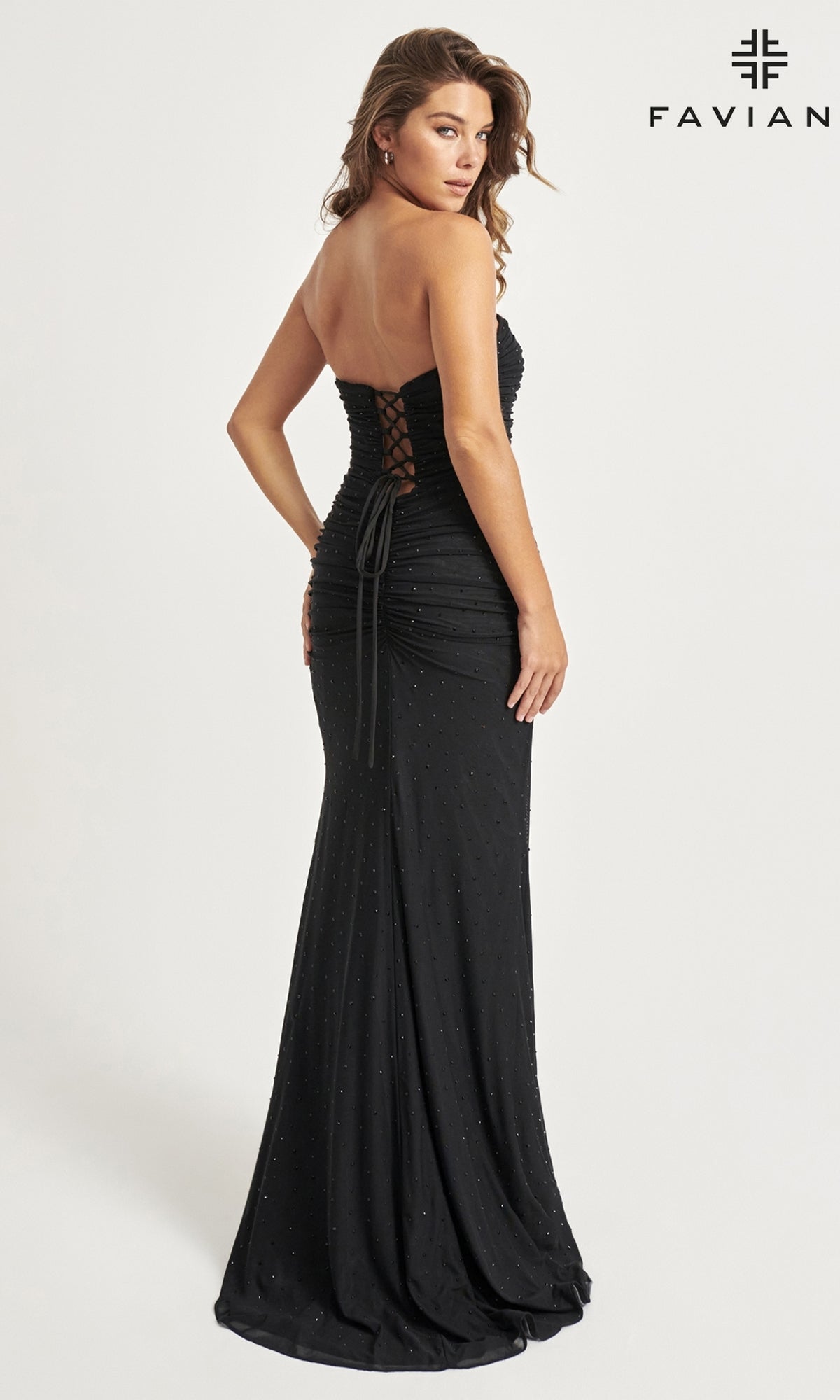 A woman in the Faviana Corset Long Strapless Prom Dress 11040, a floor-length black gown with a lace-up back and elegant beaded details, stands sideways and glances over her shoulder, showcasing its sparkling embellishments.