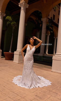 A woman in the Long Wedding Dress 11101 by Primavera stands outdoors before tall columns and palm trees, sunlight illuminating her fitted ivory gown. "PRIMAVERA" is displayed beside her.
