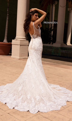 A woman stands outdoors in front of columns, facing away from the camera, wearing the Long Wedding Dress 11101 by Primavera—a fitted gown with white lace, open back, and a long train.