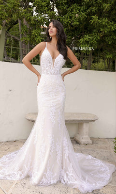 A woman in the Long Wedding Dress 11103 by Primavera, featuring an ivory mermaid silhouette with a deep v-neckline and open back, stands outdoors near a stone bench and greenery.