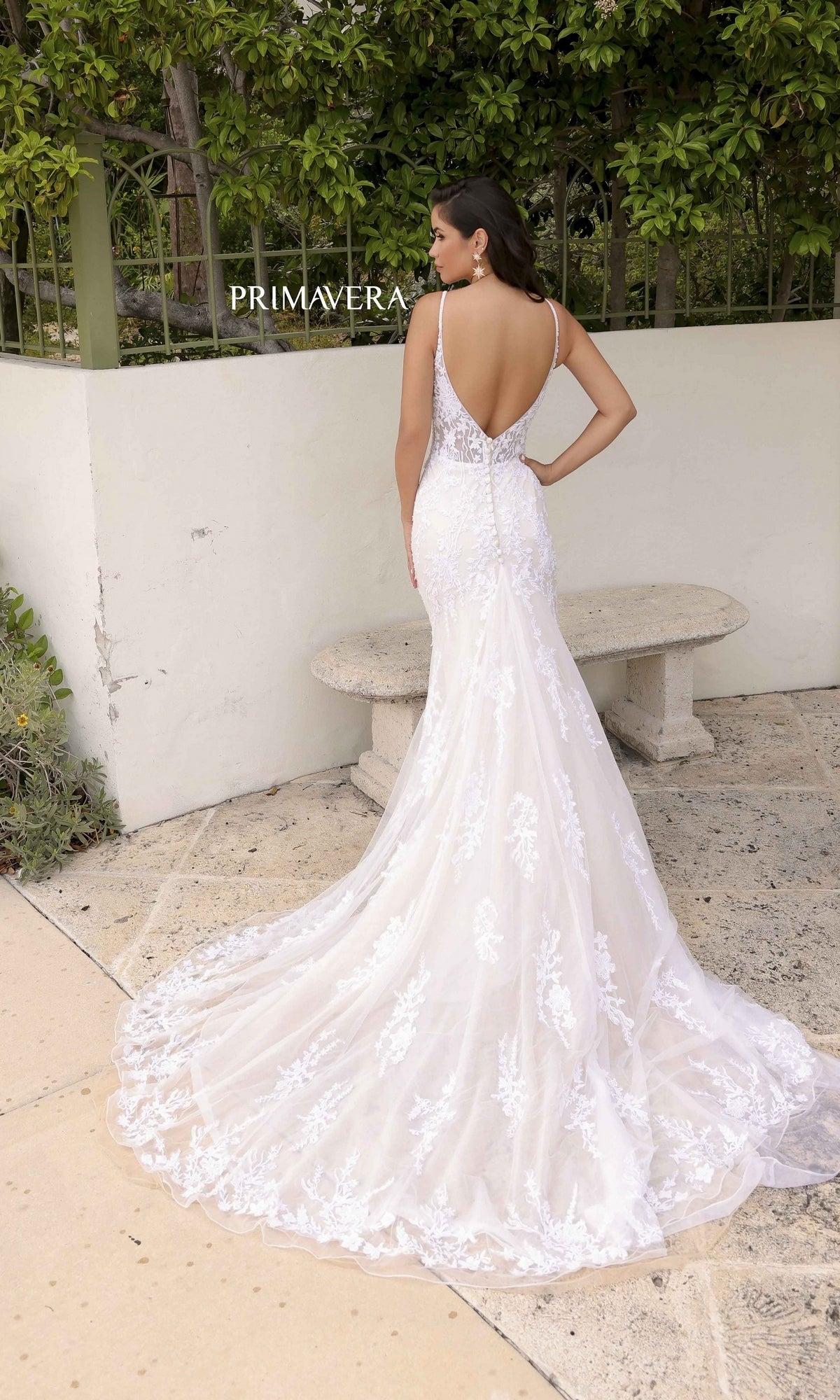 A woman stands outside by a white wall and greenery, wearing the Long Wedding Dress 11103 by Primavera—an ivory mermaid gown featuring delicate lace, a dramatic open back, and a sweeping long train.