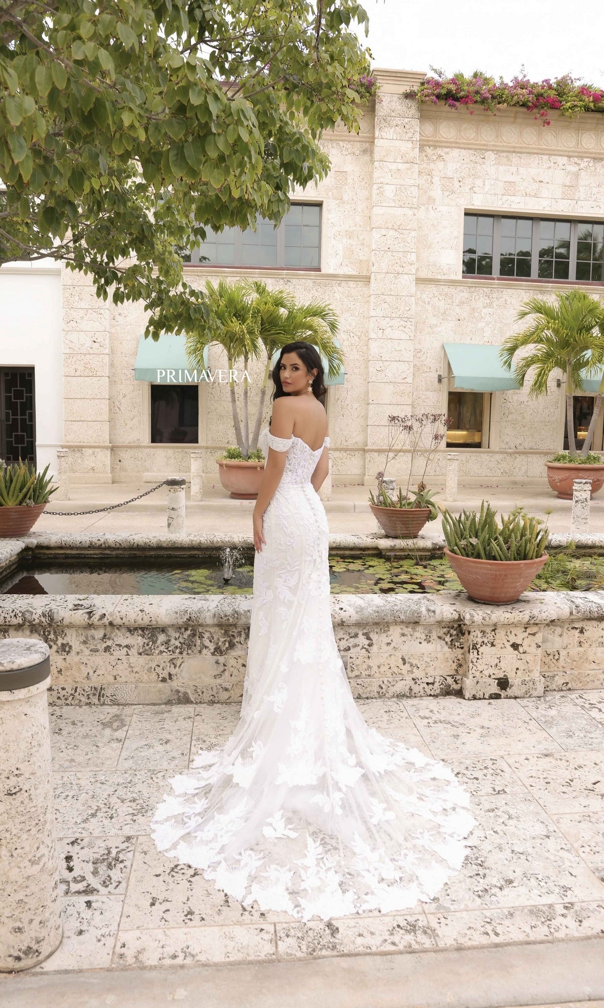 A woman in the Long Wedding Dress 11104 by Primavera, featuring off-the-shoulder lace embroidery and a long train, stands outdoors on a stone path surrounded by tropical plants and a stone building.