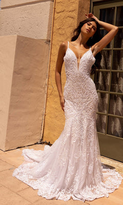 A woman stands outside by a beige wall and glass door, wearing the Long Wedding Dress 11105 by Primavera—an ivory mermaid gown with lace embroidery, thin straps, a train, and open back—her hand resting on her forehead.