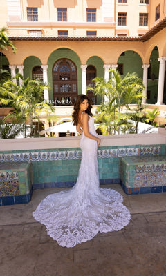 A woman wearing the Long Wedding Dress 11106 by Primavera, featuring a low v-neckline, stands before a tiled wall with "PRIMAVERA," palm trees, and a building visible in the background.