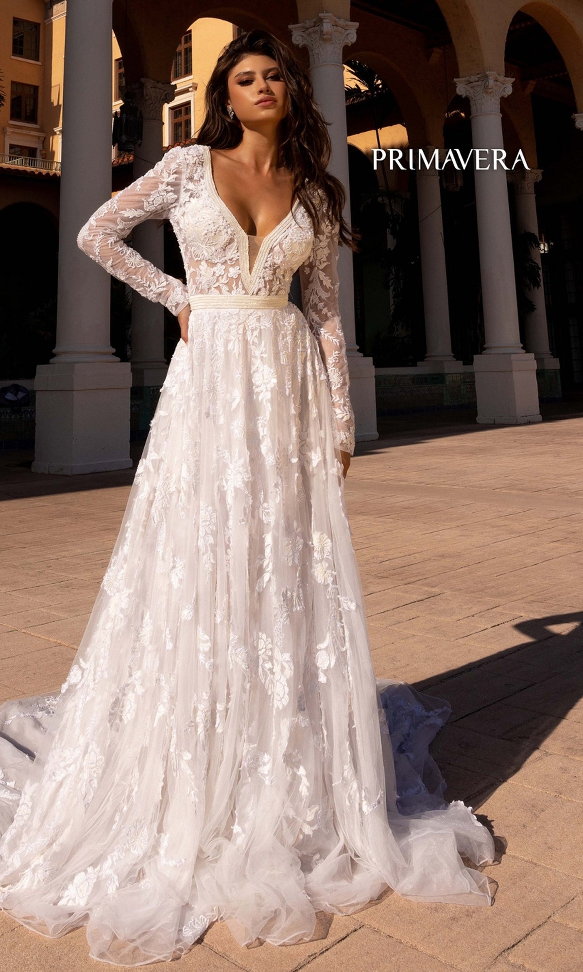 Outdoors, a woman wears the Long Wedding Dress 11111 by Primavera—an elegant ivory lace gown with long sleeves, a deep V-neckline, and floral patterns. 
