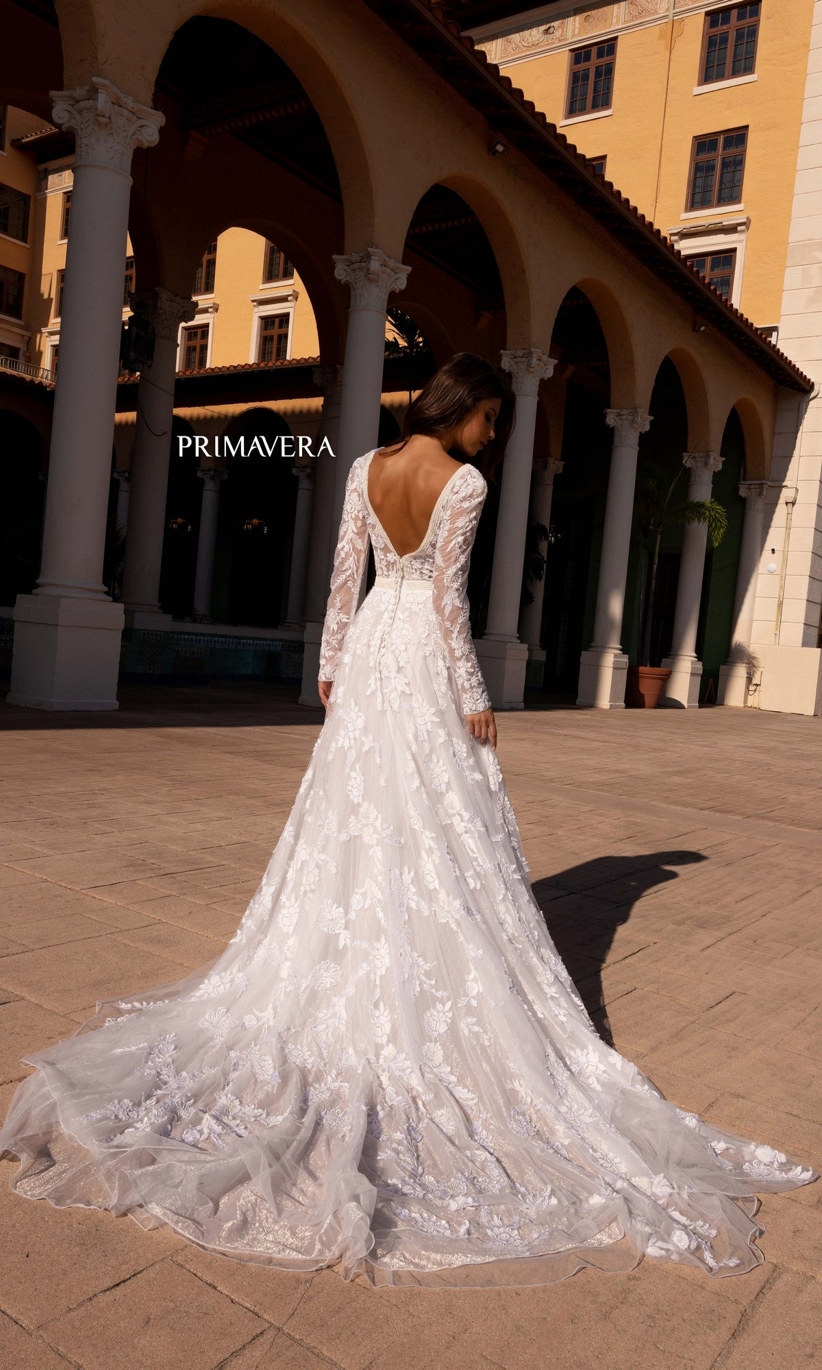 A woman stands outside facing away, wearing the Long Wedding Dress 11111 by Primavera—an ivory lace gown with long sleeves and a deep V-back—in front of a yellow building with arches and columns.