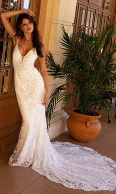 A woman models the Long Wedding Dress 11112 by Primavera, a fitted ivory lace gown with a v-neckline, sleeveless design, and long train, standing by a potted plant and tan building.
