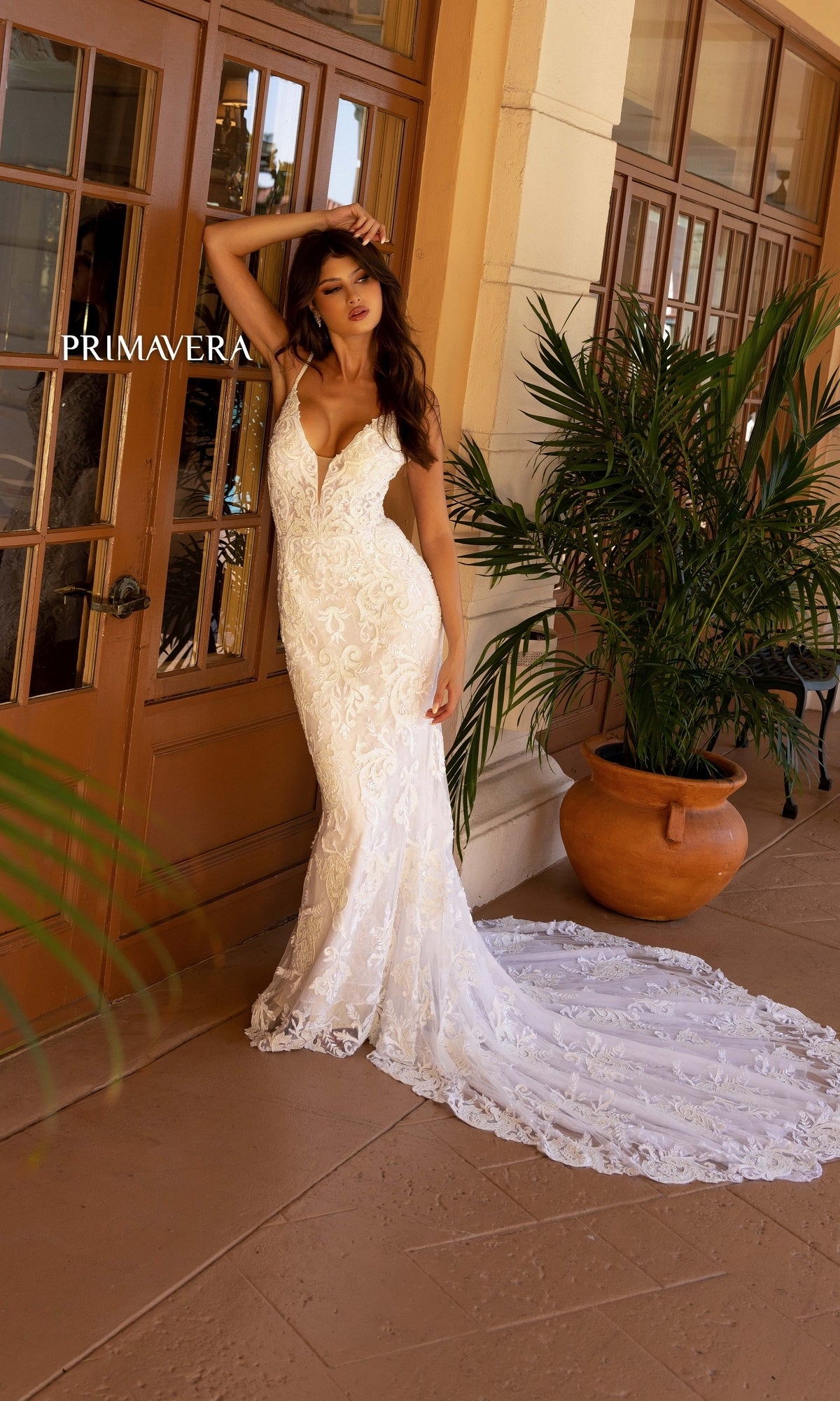 A woman in the Long Wedding Dress 11112 by Primavera, featuring a fitted ivory lace design with a v-neckline and long train, stands by glass doors and potted plants.