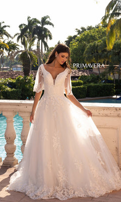 A woman wearing the Long Wedding Dress 11134 by Primavera, an ivory A-line gown with cape short sleeves, stands by a poolside balustrade amid palm trees and greenery. "PRIMAVERA" is visible in the image.